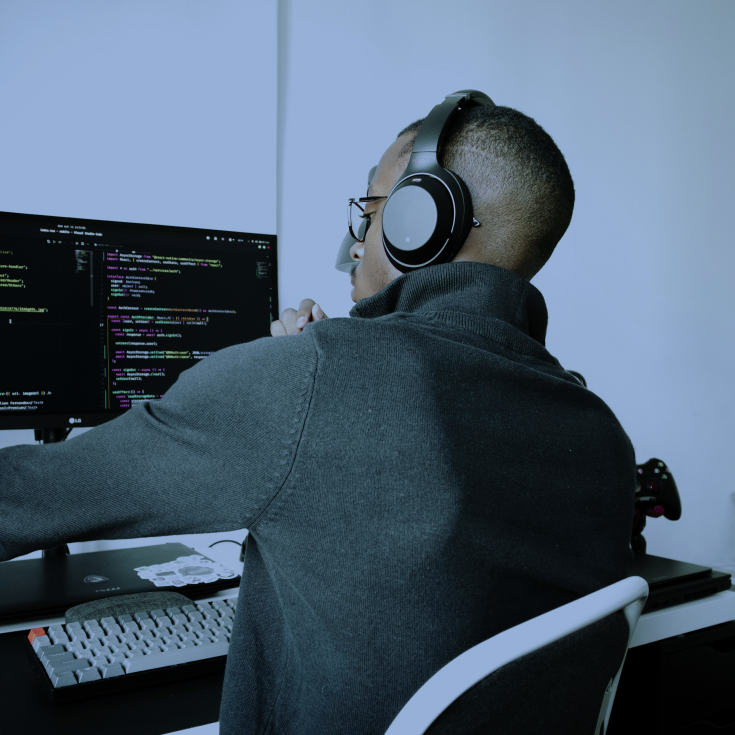 A person wearing headphones and glasses, sitting at a desk, looking at a computer screen with code on it