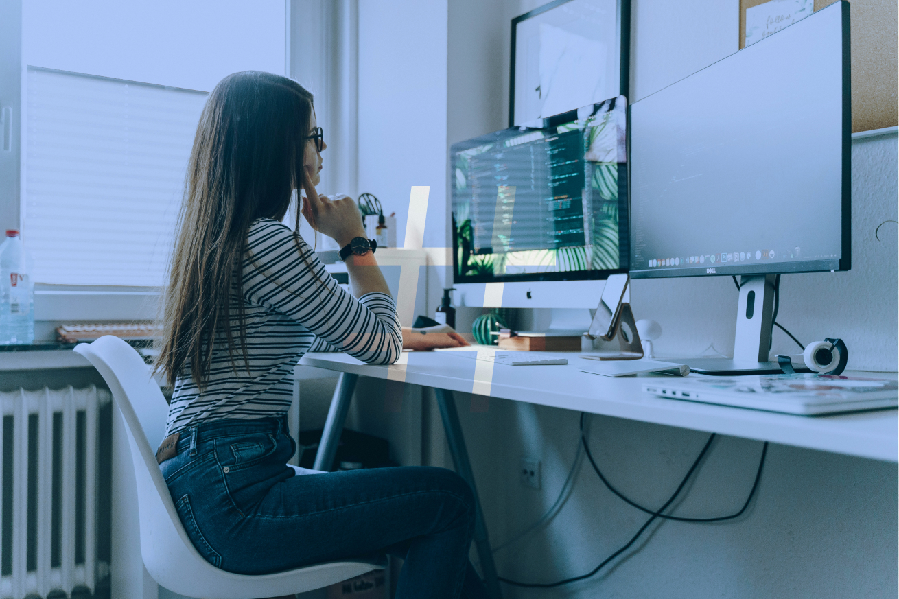 A person sits at a desk with two computer monitors displaying code