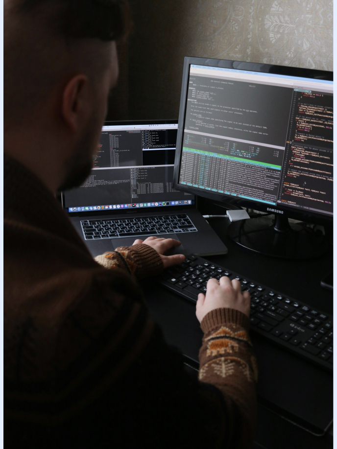 A person is seated at a desk, working on a computer with multiple monitors displaying code